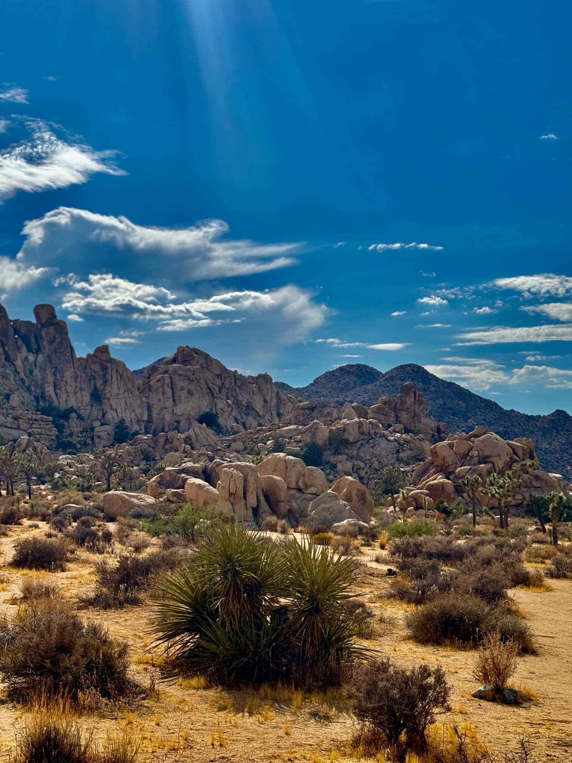 Sunbeams breaking through clouds over a boulder-strewn ridge and distant mountains in Joshua Tree
